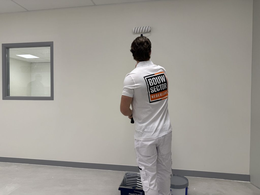 Schilder in wit poloshirt rolt een lichte wand naast een binnenraam met grijs kozijn