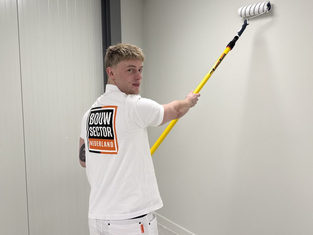 Jonge schilder kijkt om terwijl hij met een verlengsteel een witte muur saust