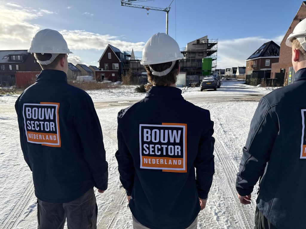 Drie vakmensen met witte helm en bedrijfsjas kijken over een besneeuwde straat met nieuwe woningen