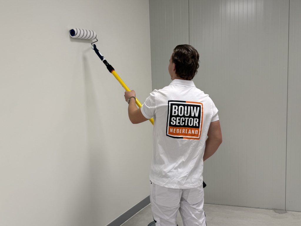 Schilder van Bouwsector Nederland zet een witte binnenwand in de verf met een roller aan een telescoopsteel